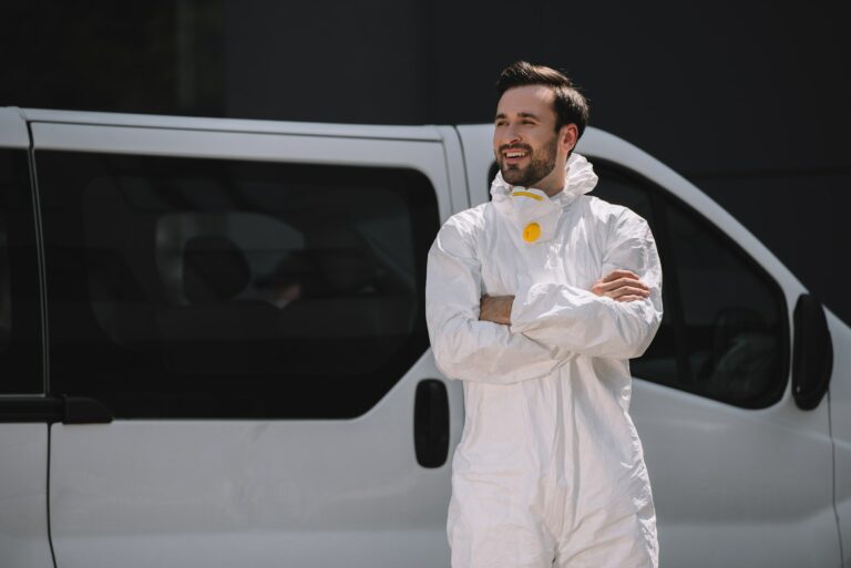 smiling pest control worker in uniform standing with crossed arms near car on street